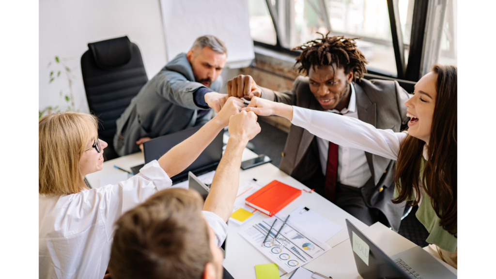 Diverse group of professionals sitting at a table celebrating with fist bumps, symbolizing thought diversity and team collaboration